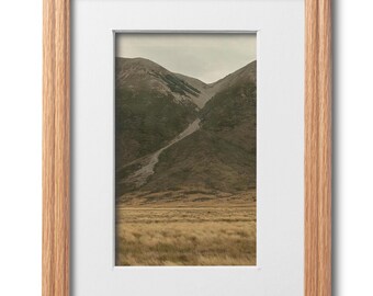 May include: A framed landscape photograph featuring a mountain range with a valley. The photograph is set in a light brown wooden frame with a white mat. The mountain slopes are covered in green vegetation and the foreground shows golden-brown grass.
