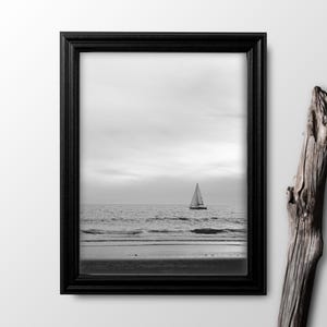 May include: A black and white photograph of a sailboat on the ocean with a sandy beach in the foreground. The photo is framed in a black frame.