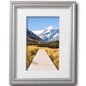 May include: A wooden walkway leads towards a snow-capped mountain peak in a field of tall grass. The sky is blue with white clouds.