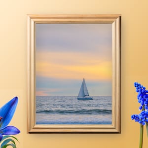 May include: A framed photograph of a sailboat on the ocean at sunset. The sailboat is white and has a white sail. The ocean is blue and the sky is a soft pink and orange.