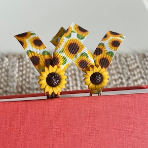 May include: Two sunflower-patterned ribbon bookmarks with glitter accents. The bookmarks are attached to a red book.