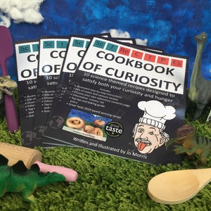 May include: A stack of four black and white cookbooks with the title "Cookbook of Curiosity" and a cartoon chef with a white chef's hat and a tongue sticking out. The books are surrounded by toy dinosaurs, a rolling pin, a wooden spoon, and a grater.