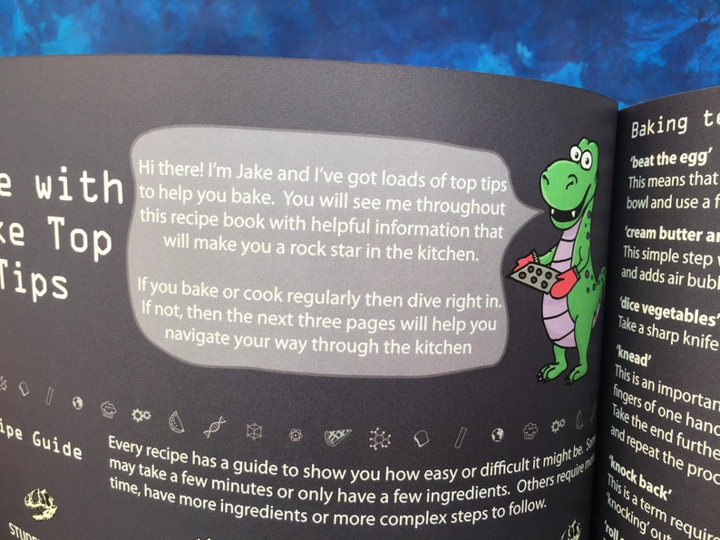 May include: A green dinosaur character wearing red oven mitts holds a baking sheet with chocolate chip cookies. The text reads "Hi there! I'm Jake and I've got loads of top tips to help you bake. You will see me throughout this recipe book with helpful information that will make you a rock star in the kitchen."  The text also reads "Every recipe has a guide to show you how easy or difficult it might be, some may take a few minutes or only have a few ingredients. Others require time, have more ingredients or more complex steps to follow."