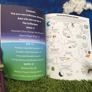 May include: A colorful illustrated book with a blue, green, and pink background. The book features a map with a choose your own adventure theme. The map includes a variety of recipes with names like "Herschel's Choc-Orange Star Biscuits" and "Turing's Banana Split Bombe Glac&eacute;e".