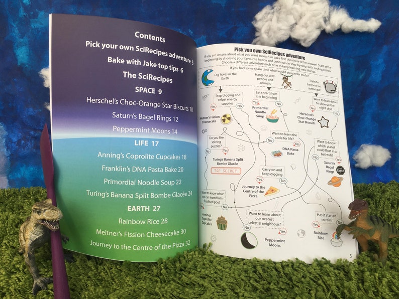 May include: A colorful illustrated book with a blue, green, and pink background. The book features a map with a choose your own adventure theme. The map includes a variety of recipes with names like "Herschel's Choc-Orange Star Biscuits" and "Turing's Banana Split Bombe Glac&eacute;e".