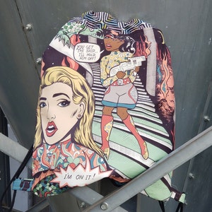 May include: A colorful drawstring bag with a comic book style design featuring two women. One woman is blonde with a red and green patterned shirt and the other woman has brown skin and is wearing a yellow shirt and grey shorts. The bag has the text "You get the ship... I'll hold him off!" and "I'm on it!" printed on it.