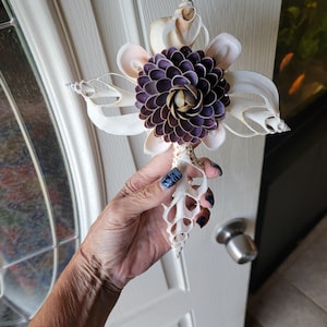 May include: A decorative cross ornament crafted from various white seashells, featuring a central flower-like cluster of small, dark purple shells. The structure is composed of elongated, spiraled shells arranged in a cross shape with a textured finish.