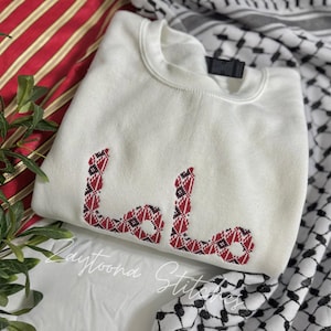 May include: A white sweatshirt with red and black embroidered Arabic script. The script is in a geometric pattern. The sweatshirt is folded and rests on a red and white striped fabric and a black and white checkered scarf.