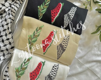 May include: Three white, beige, and black sweatshirts with an embroidered design of a watermelon shaped like the outline of Palestine, a green olive branch, and a black and white keffiyeh.