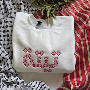May include: A white crewneck sweatshirt with red and black embroidered Arabic script. The sweatshirt is folded and displayed on a patterned fabric background. The text "Moonay Stitches" is visible in the background.