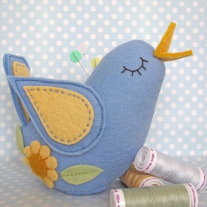 May include: A blue felt bird pincushion with yellow beak and wings, a yellow flower, and green leaves. The bird is sitting on a spool of thread and two spools of thread are in front of it.