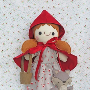 Op de afbeelding: Een pop met een rode kap, bruin haar, een grijze en rode jurk en een bruine mand. De pop houdt een grijs wolvenknuffel vast. Er liggen koekjes met roze en witte sprinkles op de vloer.