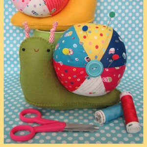 Könnte beinhalten: Zwei bunte Nadelkissen in Form von Schnecken. Die größere Schnecke ist gelb mit einer mehrfarbigen Patchwork-Schale. Die kleinere Schnecke ist grün mit einer blauen, gelben und roten Patchwork-Schale. Beide Schnecken haben schwarze Augen und einen lächelnden Mund.