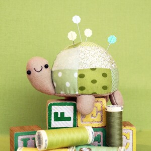 Pode incluir: Um alfineteiro de tartaruga de tecido patchwork verde e branco com um rosto sorridente está sentado em cima de blocos de madeira que soletram a letra