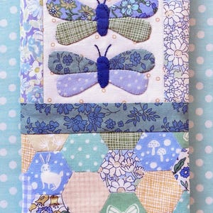 Könnte beinhalten: Ein handgefertigtes Stoffstück mit zwei blauen Schmetterlingsapplikationen auf cremefarbenem Hintergrund. Das Design integriert Blumen- und geometrische Muster in Blau-, Grün- und Cremetönen. Es handelt sich wahrscheinlich um ein kleines Quilt oder Dekorationsobjekt.