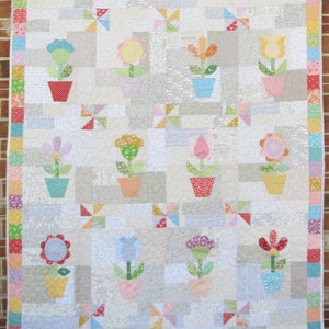 May include: A white quilt with a patchwork design featuring colorful flowers in pots. The quilt has a border of pink, green, blue, and orange squares.