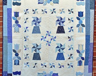 Könnte beinhalten: Eine weiße Bettdecke mit einem blauen und weißen Blumenmuster. Die Bettdecke zeigt ein sich wiederholendes Muster aus Windmühlen und Tulpen. Die Bettdecke ist mit einem blauen und weißen gemusterten Stoff eingefasst.