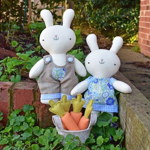 May include: Two white plush bunny dolls with embroidered faces, one wearing brown overalls with a floral pocket and the other a blue floral dress. A felt basket holds orange and green carrot toys, all set against a garden backdrop.