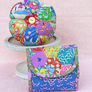 以下が含まれることがあります： 中央にボタンが付いたカラフルなパッチワークの針山と、青い裏地が付いた同じパッチワークの布製のポーチ