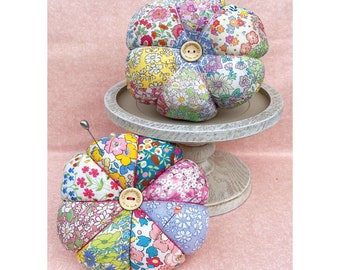 May include: Two floral pincushions in the shape of a flower. The pincushions are made of fabric with a variety of floral patterns. The pincushions are sitting on a white pedestal. The pincushions have a button in the center with the words "Cottonwood Creek" on them.