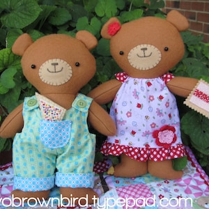 Puede incluir: Dos osos de peluche marrones que llevan ropa. El oso de la izquierda lleva un mono azul con un estampado verde y blanco. El oso de la derecha lleva un vestido rosa y blanco con un ribete rojo y blanco de cuadros.