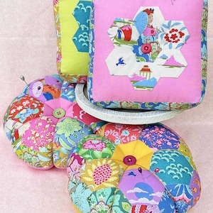 May include: A selection of handmade pincushions in various shapes and sizes. The pincushions display vibrant floral and geometric patterns in pink, green, blue, and yellow. Some have a button in the centre and pins inserted. The pincushions are arranged on a pink surface.