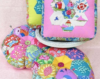 May include: A collection of colorful, handmade pincushions. The pincushions feature floral and geometric patterns in shades of pink, green, blue, and yellow. Some have a button in the center and pins inserted. The pincushions are arranged on a pink surface.