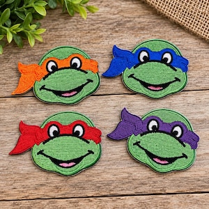 May include: Four embroidered patches featuring the faces of green turtles. Each turtle has a wide smile and wears a colored eye mask: one orange, one blue, one red, and one purple. The patches are circular with detailed stitching.