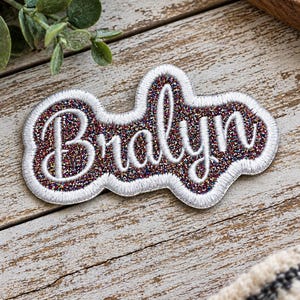 May include: A custom embroidered patch featuring the name Bralyn in white cursive script. The lettering is set against a dark, multi-colored glitter background, surrounded by a thick, white stitched border that follows the shape of the text.