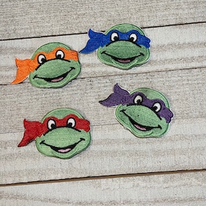 May include: Four embroidered patches featuring the Teenage Mutant Ninja Turtles. Each patch depicts a turtle head with a different colored mask: orange, blue, red, and purple. The patches are green with black eyes and smiling mouths.