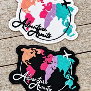 May include: Two embroidered patches featuring a world map design with continents in peach, pink, purple, and teal. One patch has a white background and black text, the other a black background and white text. Both read Adventure Awaits.