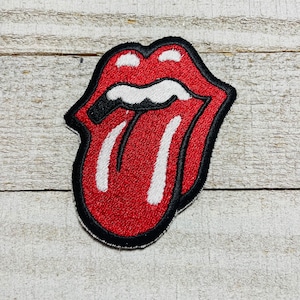 May include: An embroidered patch featuring a stylized red mouth with a protruding white tongue, outlined in black thread. The design depicts a pair of lips with white teeth and a prominent tongue, creating a bold graphic emblem with a textured finish.
