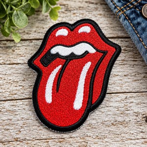 May include: An embroidered patch featuring the iconic Rolling Stones tongue and lips logo. The design consists of a bright red tongue and lips outlined in black, with white accents on the teeth and tongue, creating a bold graphic appearance.