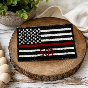 May include: A rectangular embroidered patch featuring a black and white American flag design with a horizontal red stripe running through the center. The number 501 is stitched in red thread over the lower portion of the flag design.