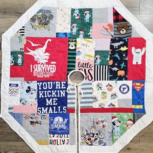 May include: A multi-colored patchwork Christmas tree skirt featuring various graphic prints including dinosaurs, cartoon characters, and text such as I SURVIVED JURASSIC QUEST, YOU'RE KICKIN' ME SMALLS, and BIG JOEL'S SAFARI, finished with a white border and ties.