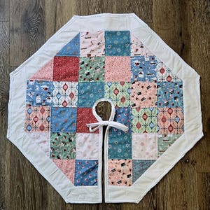 May include: A quilted Christmas tree skirt with a patchwork design. The skirt features squares of red, pink, blue, and green fabric with snowflake and snowman patterns. A white border and tie closure complete the design.