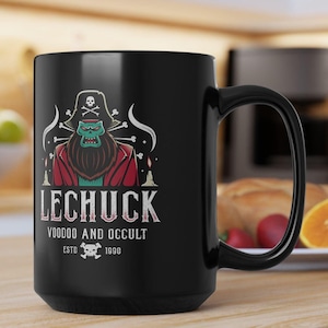 May include: Black ceramic mug featuring a cartoon illustration of a pirate with the text "LECHUCK VOODOO AND OCCULT ESTD 1990". The pirate has a green face, a long beard, and wears a red coat and a pirate hat with a skull and crossbones.