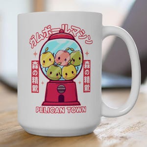 Könnte beinhalten: Weiße Keramik-Tasse mit einem roten Kaugummiautomaten-Design und Cartoon-Fruchtfiguren. Die Tasse hat japanischen Text und die Worte "Pelican Town" aufgedruckt. Die Tasse steht auf einer Holzoberfläche.