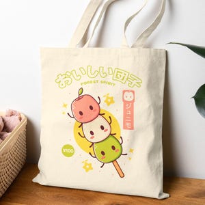 May include: Cream tote bag with a colourful illustration of three mochi characters on a stick. The design includes Japanese text and a yellow circle with a price tag. The bag has long handles.
