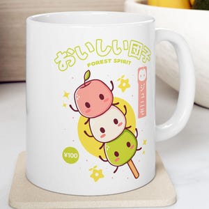 Könnte beinhalten: Weiße Keramik-Tasse mit einer farbenfrohen Illustration von drei Mochi-Bällchen auf einem Stock. Das Design enthält japanischen Text und ein ¥100-Symbol. Die Tasse hat einen gebogenen Griff und steht auf einem beigen Untersetzer.