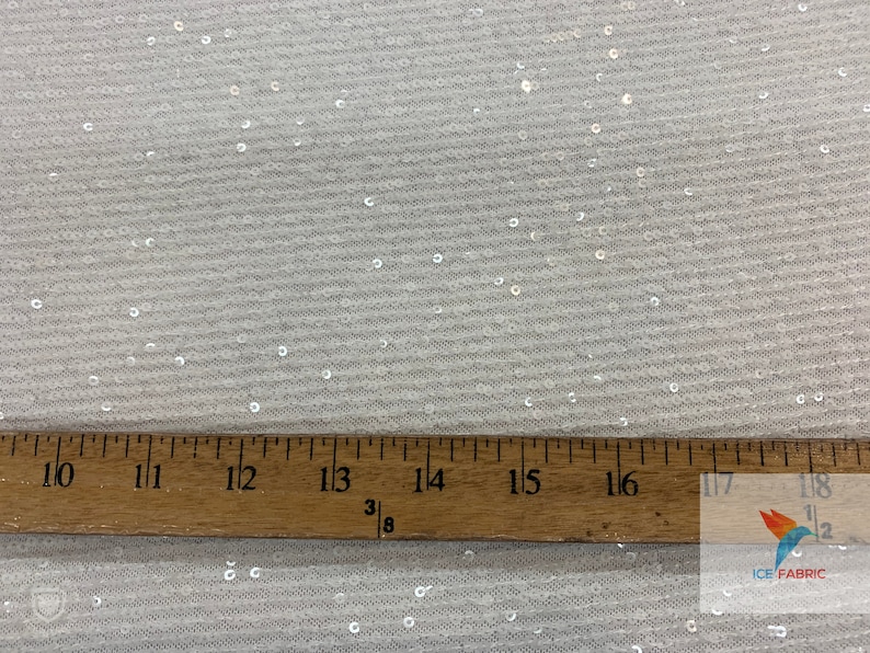 May include: White fabric with small, iridescent sequins. A ruler is placed on the fabric, showing measurements from 10 to 18 inches.