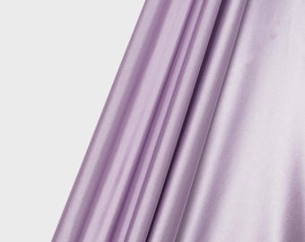 May include: A length of light purple satin fabric with a smooth, reflective surface and a subtle sheen. The material is draped in soft, vertical folds, creating a series of highlights and shadows across the fabric. A logo reads ICE FABRICS.