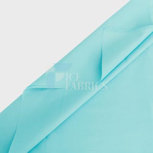 May include: A solid light turquoise fabric folded diagonally to reveal a smooth, matte texture. The fabric features a watermark logo in the centre that reads ICE FABRICS in a clean, sans-serif font, layered over a semi-transparent blue graphic.