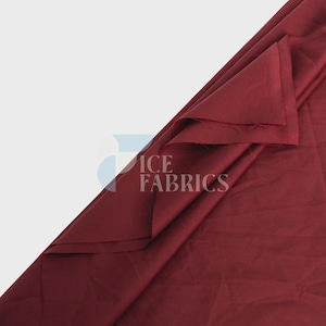 May include: A solid deep burgundy fabric draped to show its texture and folds. The material has a smooth, matte finish with a consistent dark red color throughout. A watermark with the text ICE FABRICS is visible on the surface of the fabric.
