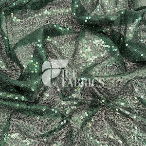 May include: Close-up of a dark green sequin fabric. The fabric is densely covered in small, round sequins that catch the light, creating a shimmering effect. The fabric is slightly ruffled, adding texture. The text "ICE FABRICS" is visible.