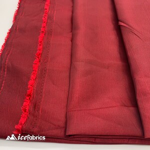 May include: Close-up of a deep red fabric with a subtle sheen. The fabric is folded and has a fringed edge.