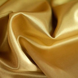 Op de afbeelding: Een close-up van een gladde, gouden satijnen stof met zachte plooien en kreukels. De stof heeft een luxe en elegante uitstraling.