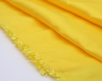 May include: A length of bright yellow fabric with a smooth, solid-colored surface and a visible frayed edge. The material is folded in soft, cascading layers, showcasing the vibrant, saturated yellow hue and the texture of the textile weave.