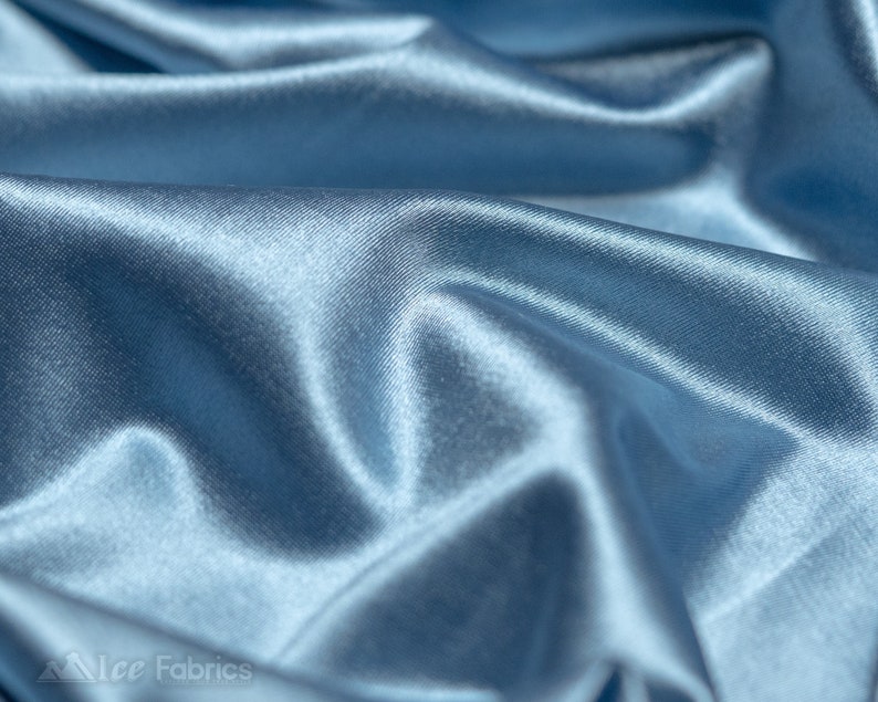 Peut inclure: Un tissu satin&eacute; bleu clair avec une surface lisse et r&eacute;fl&eacute;chissante dot&eacute;e d'un &eacute;clat subtil. Le mat&eacute;riau est drap&eacute; en plis doux et fluides qui cr&eacute;ent des ombres profondes et des reflets lumineux sur le textile. Le texte Ice Fabrics est visible.