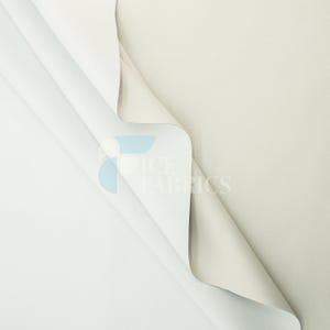 Puede incluir: Una pieza doblada de tela color blanco hueso con una textura lisa y mate. El material está drapeado para revelar ambos lados, mostrando un color crema claro consistente. Un logotipo azul y blanco muestra el texto ICE FABRICS.